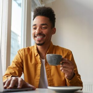 african-american-sitting-table-cafe-working-laptop-wears-yellow-shirt-drinks-aromatic-coffee-communicates-with-his-sister-which-is-far-another-country-enjoys-work 2